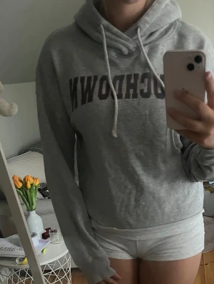 Grå hoodie med TOUCHDOWN-tryck - Säljer en grå hoodie med texten 'TOUCHDOWN' i svart på bröstet. Hoodien har huva med vita dragsnören och är croppad i modellen. Perfekt för en avslappnad och sportig stil. Materialet är mjukt och skönt, troligen bomullsblandning.