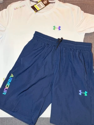 Under Armour träningsset t-shirt & shorts - Säljer ett sportigt träningsset från Under Armour med en vit t-shirt och marinblå shorts. Båda plaggen har logga och text i regnbågsfärger. T-shirten är rundhalsad och shortsen har resårmidja med dragsko. Perfekt för gym eller löpning.