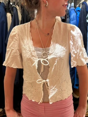 Vit blus med broderi och knyt - Söt vit blus med broderade detaljer och två knyt framtill. Blusen har korta ärmar, vågig kant och är tillverkad i ett lätt bomullstyg🧘🏼‍♀️🤍