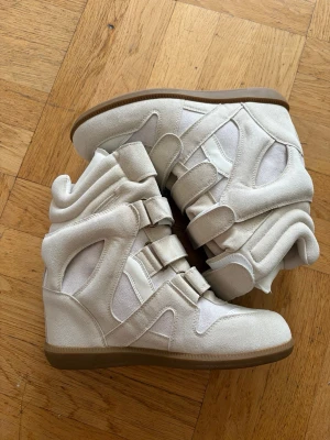 Isabel marant - Snygga vita sneakers med högt skaft och kilklack. Skorna har flera kardborreband och är gjorda i en mix av mockaimitation och textil. Yttersulan är i gummi och designen är chunky och modern, perfekt för en trendig streetstyle-look.