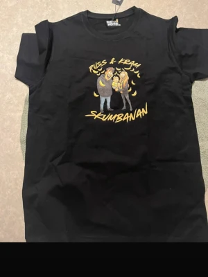 Svart t-shirt med skumbanan-tryck - Svart t-shirt med färgglatt tryck av två personer och texten 'Russ & Kram Skumbanan' på bröstet. T-shirten har rund hals och korta ärmar. Perfekt för dig som gillar roliga motiv och vill sticka ut lite extra.
