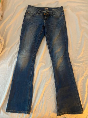 Blå bootcut jeans från ONLY - Säljer ett par blå bootcut jeans från ONLY med snygga slitningar och klassisk femficksmodell. Jeansen har normal midja och är gjorda i ett mjukt denimtyg med lite stretch. Perfekta för dig som gillar en avslappnad men trendig look.