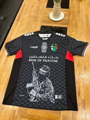 Svart fotbollströja Palestina Capelli - Säljer en svart fotbollströja från Capelli med Palestinas klubbmärke, Bank of Palestine-tryck och grafiskt motiv framtill. Tröjan har röda och gröna detaljer på ärmarna, korta ärmar och är tillverkad i ett lätt, ventilerande material.
