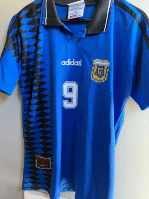 Argentina retro fotbollströja blå - Säljer en blå retro Argentina fotbollströja från Adidas med svart krage, vita detaljer och nummer 9. Tryck med 'Batistuta' på ryggen och AFA-emblem på bröstet. Klassisk design med svarta trianglar längs sidan. Perfekt för fotbollsfans!