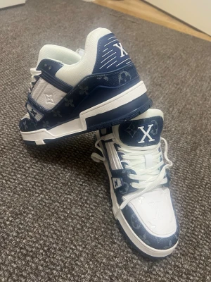 Blåvita sneakers med monogramdetaljer - Säljer ett par riktigt snygga sneakers i blått och vitt med tydliga monogramdetaljer och X-logga på plösen. Skorna har vita snören, vadderad krage och en chunky sula. Materialmix av textil och syntet ger en modern vibe. Perfekta för dig som vill sticka ut.