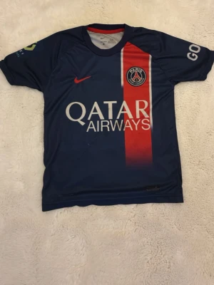PSG matchtröja Mbappé nr 7 - Säljer en blå Paris Saint-Germain matchtröja med röd vertikal rand, Qatar Airways-tryck och PSG-logga på bröstet. På ryggen står det Mbappé och nummer 7. Perfekt för fotbollsfans! Kortärmad och tillverkad i lätt funktionsmaterial.