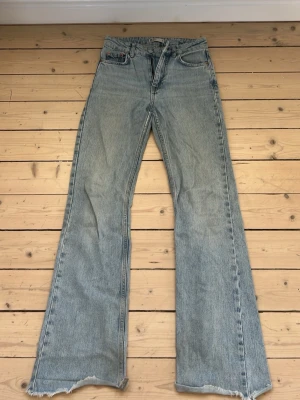 Ljusblå utsvängda jeans med rå kant - Säljer ett par ljusblå jeans med utsvängda ben och råa, fransiga benslut. Klassisk femficksmodell i denim med hög midja och dragkedja fram. Perfekta för en avslappnad och trendig look.