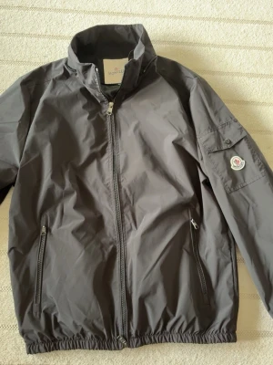Svart vindjacka från Moncler - Snygg svart vindjacka från Moncler med dragkedja framtill, två sidofickor med dragkedja och en ficka på ärmen med Moncler-logga. Jackan har huva och elastiska muddar vid ärmslut och nederkant. Perfekt för blåsiga dagar.