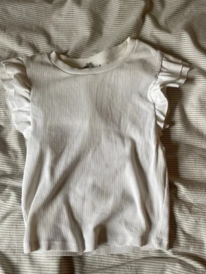 Topp från Zara - Supersöt vit ribbad topp från Zara med korta volangärmar. Perfekt för dig som gillar en enkel men ändå söt stil. T-shirten har rund halsringning och är gjord i ett mjukt material som känns skönt mot huden.