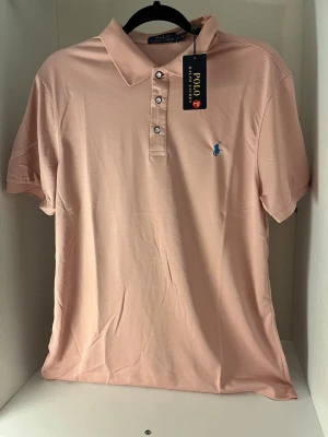 Ljusrosa pikétröja från Polo Ralph Lauren - Snygg ljusrosa pikétröja från Polo Ralph Lauren med klassisk krage, korta ärmar och knappar framtill. Tröjan har en broderad blå logga på bröstet och är tillverkad i mjuk bomull. Perfekt för en stilren och avslappnad look.