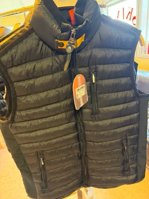 Svart dunväst med hög krage - Snygg svart dunväst med quiltad design och hög krage. Västen har dragkedja framtill, tre praktiska fickor med dragkedja och detaljer i gult vid halsen. Perfekt för lager-på-lager och håller dig varm under kyliga dagar.