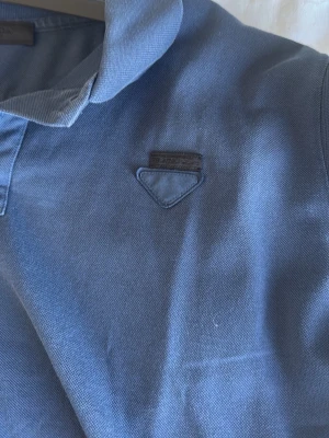 Blå pikétröja med krage från Prada  - Snygg blå pikétröja med klassisk krage och korta ärmar. Tröjan har en liten detalj på bröstet och är tillverkad i ett mjukt bomullsmaterial. Perfekt för en avslappnad och stilren look.
