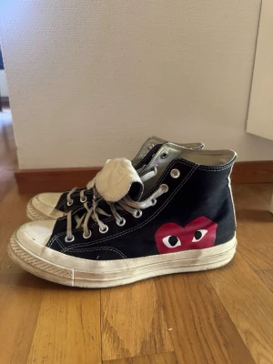 CONVERSE CDG - Svarta high top sneakers med vit sula och vita snören. På sidan finns en röd hjärtdetalj med ögon, vilket ger skorna en unik och lekfull look. Tillverkade i canvasmaterial med klassisk gummisula och metallöljetter för snörningen.