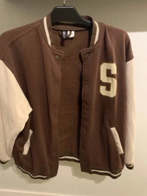 Brun och beige bomberjacka Seattle - Snygg brun bomberjacka med beige ärmar och detaljer. Stor S på bröstet och texten 'Seattle' i vitt på ryggen. Jackan har ribbade muddar, tryckknappar och två fickor framtill. Perfekt för en sportig och avslappnad stil.
