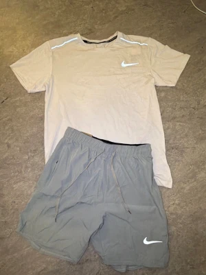 Nike set: ljus t-shirt & grå shorts - Säljer ett sportigt set från Nike med en ljus t-shirt och ett par grå shorts. T-shirten har rund hals och Nike-logga på bröstet, medan shortsen har dragsko i midjan och logga på benet. Perfekt för träning eller chill dagar.