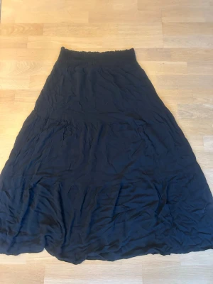 Svart långkjol - Svart långkjol med bred smock i midjan som ger en bekväm passform. Kjolen har en lätt utställd form och är gjord i ett tunt, mjukt tyg som faller snyggt. Perfekt för dig som gillar en enkel och stilren look.
