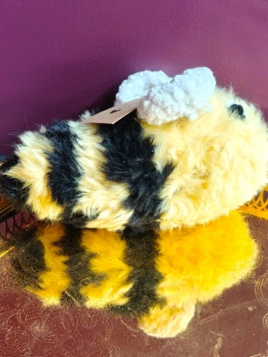 Fluffigt bi-gosedjur i plysch - Säljer ett supermjukt gosedjur i form av ett bi. Biet har gul och svart randig kropp, vita fluffiga vingar och broderade ögon. Tillverkat i plyschmaterial och har en rund, mysig form som gör det perfekt som dekoration eller kramkompis.