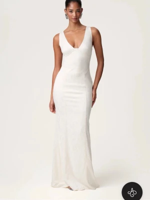 Vit ”ANDRIA DRESS” från Adoore - Elegant vit långklänning med djup v-ringning och ärmlös design från Adoore. Klänningen har en figurnära passform och är tillverkad i ett material som ger en lätt glansig yta. Perfekt för speciella tillfällen där du vill känna dig extra fin. Klänningen har använts en gång innan