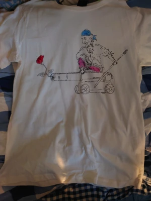 Vit t-shirt med piratmotiv - Säljer en vit t-shirt med ett unikt tryck på ryggen som föreställer en pirat med blå bandana och rosa byxor sittandes på en kanon med en röd ros i mynningen. T-shirten har korta ärmar och är gjord i mjuk bomull. Perfekt för dig som gillar coola och annorlunda motiv.