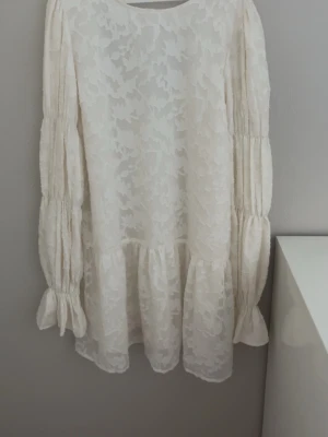 Vit kort klänning med volang - Superfin krämig vit kort klänning från Gina tricot med volangkant och långa ärmar med knyt vid ärmslut. Klänningen har ett subtilt blommigt mönster i tyget och en öppen rygg. Perfekt för sommarens alla tillfällen!🎀