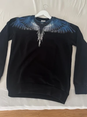 Svart sweatshirt Marcelo Burlon - En Marcelo Burlon swestshirt i mycket bra skick. Storlek XS (passar S). Äkta såklart (kvitto finns). Svart sweatshirt från Marcelo Burlon County of Milan med ikoniskt blått och vitt vingmönster över axlar och bröst. Tröjan har rund halsringning och ribbade muddar. Perfekt för dig som vill sticka ut med en unik design.