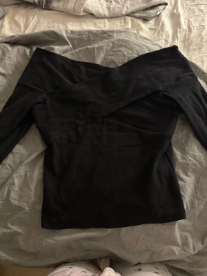 Svart offshoulder topp - Säljer en svart offshoulder topp med lång ärm och snygg omlottdesign upptill. Toppen har en tight passform och är perfekt för dig som vill ha en stilren men ändå trendig look. Materialet känns mjukt och stretchigt, vilket gör den bekväm att bära.