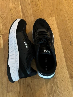 Svarta BOSS sneakers med vit sula - Snygga svarta sneakers från BOSS med vit, räfflad sula och ventilerande meshdetaljer. Skorna har klassisk låg profil, svarta skosnören och diskret BOSS-logga på sidan. Perfekta för en trendig och sportig look. Pris kan diskuteras vid snabb affär!