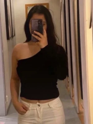 Svart oneshoulder topp - Trendig svart oneshoulder topp med lång ärm på ena sidan och axelbandslös på andra. Sitter tight och är croppad, perfekt till höga jeans eller kjol. Enkel och stilren design som passar till många olika looks.
