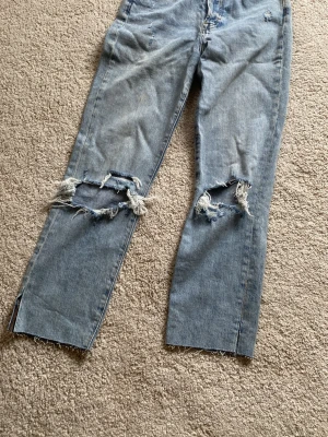 Ljusblå slitna raka jeans - Säljer ett par ljusblå jeans med raka ben och stora slitningar vid knäna. Jeansen har råa, avklippta benslut och är gjorda i klassisk denim. Perfekta för en avslappnad och trendig look.( Titta noga på bilder)