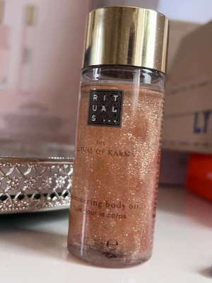Rituals The Ritual of Karma Shimmering Body Oil - Glittrande kroppsolja från Rituals i serien The Ritual of Karma. Oljan har en skimrande, guldig färg och kommer i en genomskinlig flaska med guldfärgat lock. Perfekt för att ge huden en snygg glow och lyster.