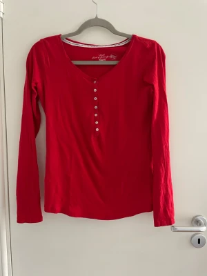 Röd långärmad topp med knappar - Snygg röd långärmad topp med v-ringning och dekorativa knappar framtill. Tillverkad i mjuk bomull som känns skön mot huden. Perfekt att styla med jeans eller kjol för en enkel men trendig look.