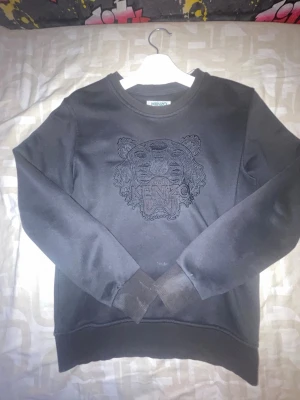 Svart sweatshirt från Kenzo med tiger - Cool svart sweatshirt från Kenzo med det ikoniska tigermotivet broderat på bröstet. Tröjan har rund halsringning, långa ärmar och ribbade muddar vid ärmslut och nederkant. Perfekt för dig som gillar streetwear och vill sticka ut.