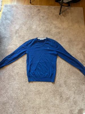 Blå stickad tröja med V-ringning - Snygg blå stickad tröja med V-ringad hals och långa ärmar. Perfekt att bära över en skjorta eller t-shirt för en clean look.