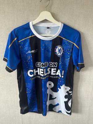 Chelsea FC blå randig matchtröja - Chelsea FC matchtröja i blått och svart med vita detaljer och tryck. Tröjan har randigt mönster, klubbmärke på bröstet och texten 'COME ON CHELSEA!' framtill. Tillverkad i lätt polyester, perfekt för fotboll eller träning.