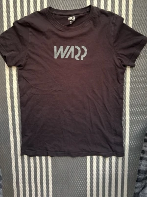 Svart t-shirt från WARP med tryck - Svart t-shirt från WARP med stort logotryck på bröstet. Klassisk rund halsringning och korta ärmar. Tillverkad i mjuk bomull som känns skön mot huden. Perfekt för en avslappnad och stilren look.