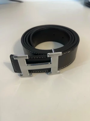 Svart skärp med H-spänne i silver - Snyggt svart skärp i skinn med ett stort H-format spänne i silverfärg. Skärpet har vita sömmar längs kanterna som ger en stilren kontrast. Perfekt accessoar för att lyfta din outfit.