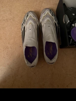 Vita och guldiga F50 fotbollsskor adidas - Använda 3 träningar men dom passar inte är för stora nypris 2200kr Säljer ett par stilrena F50 fotbollsskor i vitt med svarta detaljer och lila innersula. Sulan är guldig med vita dobbar för bra grepp på planen. Skorna har en strömlinjeformad design och är perfekta för dig som vill ha både fart och kontroll på fotbollsplanen. Väska följer med och sulor
