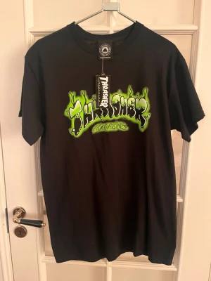 Svart Thrasher t-shirt med grön logga - Säljer en svart t-shirt från Thrasher med en stor, grön och vit grafisk logga på bröstet. Klassisk passform med rund hals och korta ärmar. Perfekt för dig som gillar streetwear och vill sticka ut med en fet logga framtill.