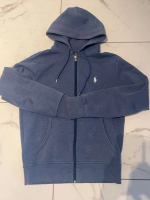 Blå zip hoodie från Polo Ralph Lauren - Snygg blå hoodie med dragkedja från Polo Ralph Lauren. Klassisk design med huva och dragsnören, två fickor framtill och den ikoniska vita loggan broderad på bröstet. Perfekt för en chill och avslappnad stil.