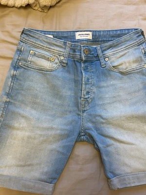 Ljusblå jeansshorts från Jack & Jones - Säljer ett par ljusblå jeansshorts från Jack & Jones med klassisk femficksdesign och uppvikta benslut. Shortsen har normal passform och är tillverkade i bomull, perfekta för varma dagar.
