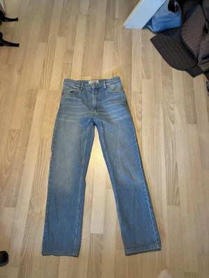 Blå raka jeans med hög midja - Säljer ett par klassiska blå jeans med rak passform och hög midja. Jeansen har fem fickor, bälteshällor och stängs med dragkedja och knapp. Materialet är kraftig denim i 100% bomull för en tidlös look. Ser ej storlek men tippar runt 27 midja och 32 längd 