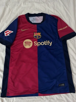Barcelona matchtröja Nike röd/blå - Säljer en officiell FC Barcelona matchtröja från Nike i rött och blått med klubbmärke, Spotify-logga och guldfärgade detaljer. Tröjan har korta ärmar, rund hals och är tillverkad i ett lätt, ventilerande funktionsmaterial. Perfekt för fotbollsträning eller att bära till vardags.