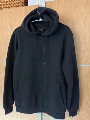 Svart hoodie från SMOG - Klassisk svart hoodie från SMOG i regular fit. Hoodien har stor huva med dragsko och en rymlig magficka framtill. Tillverkad i mjukt material som passar perfekt till chill dagar. Enkel och stilren design utan tryck eller mönster.