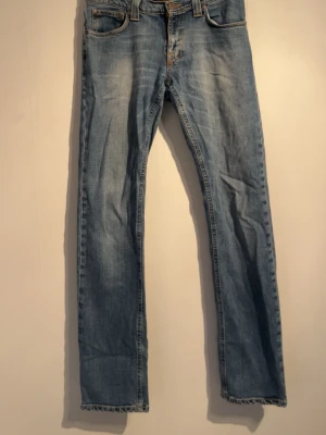 Ljusblå raka jeans från nudie - Säljer ett par ljusblå jeans med raka ben och klassisk femficksdesign. Jeansen har slitna detaljer vid benslut och bakfickor med dekorativa sömmar. Perfekta för en avslappnad och trendig look. Märket är nudie