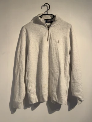 Ljusgrå halvzip från Polo Ralph Lauren - Snygg ljusgrå tröja med halv dragkedja och hög krage från Polo Ralph Lauren. Tröjan har en broderad logga på bröstet och är tillverkad i mjukt material. Perfekt för en avslappnad och stilren look.