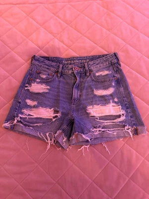 Blåa ripped jeansshorts från American Eagle - Säljer dessa super snygga blåa jeansshorts från American Eagle med slitna detaljer fram- och baktill. 