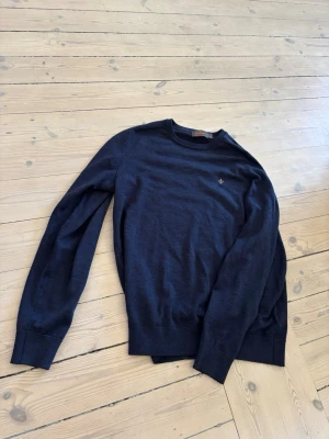 Mörkblå Morris merinoull  tröja - En stilren mörkblå merinoull tröja från Morris med rund hals och diskret broderad logga på bröstet. Tröjan har långa ärmar och ribbade muddar vid ärmslut och nederkant. Perfekt för lager på lager och enkel att matcha till olika outfits.
