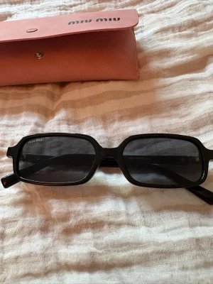 Svarta solglasögon från Miu Miu - Snygga svarta solglasögon från Miu Miu med rektangulära bågar och mörka glas. Diskret logga på sidan och medföljande rosa fodral. Perfekta för dig som vill ha en stilren och trendig accessoar till sommaren.