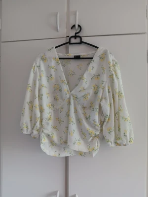 Vit blommig omlottblus med puffärm - Supersöt vit omlottblus med gula och gröna blommor. Blusen har djup v-ringning, puffiga ärmar med resår vid ärmslut och lätt rynkad midja. Tillverkad i ett tunt, strukturerat tyg som känns luftigt och somrigt. Varan är oanvänd 