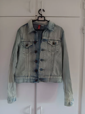Ljusblå klassisk jeansjacka - Snygg ljusblå jeansjacka med klassisk krage och bröstfickor med knapp. Jackan har bruna metallknappar framtill och på fickorna samt en lätt tvättad look. Perfekt att slänga över en hoodie eller t-shirt för en avslappnad stil.